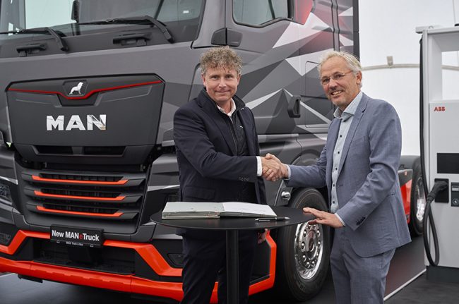 MAN Truck & Bus und ABB E-mobility verstehen sich: Im Vordergrund schütteln sich zwei Männer aus den beiden Unternehmen die Hände, im Hintergrund stehen links ein schwarzer MAN-Truck und rechts eine weiße ABB-Ladesäule.