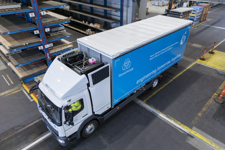 Das Foto zeigt einen LKW mit weißem Fahrerhaus und blauer Plane von oben. Die Plane trägt das Symbol von Thyssenkrupp, in der Fahrzeugfront ist das Logo der Paul Group verbaut.