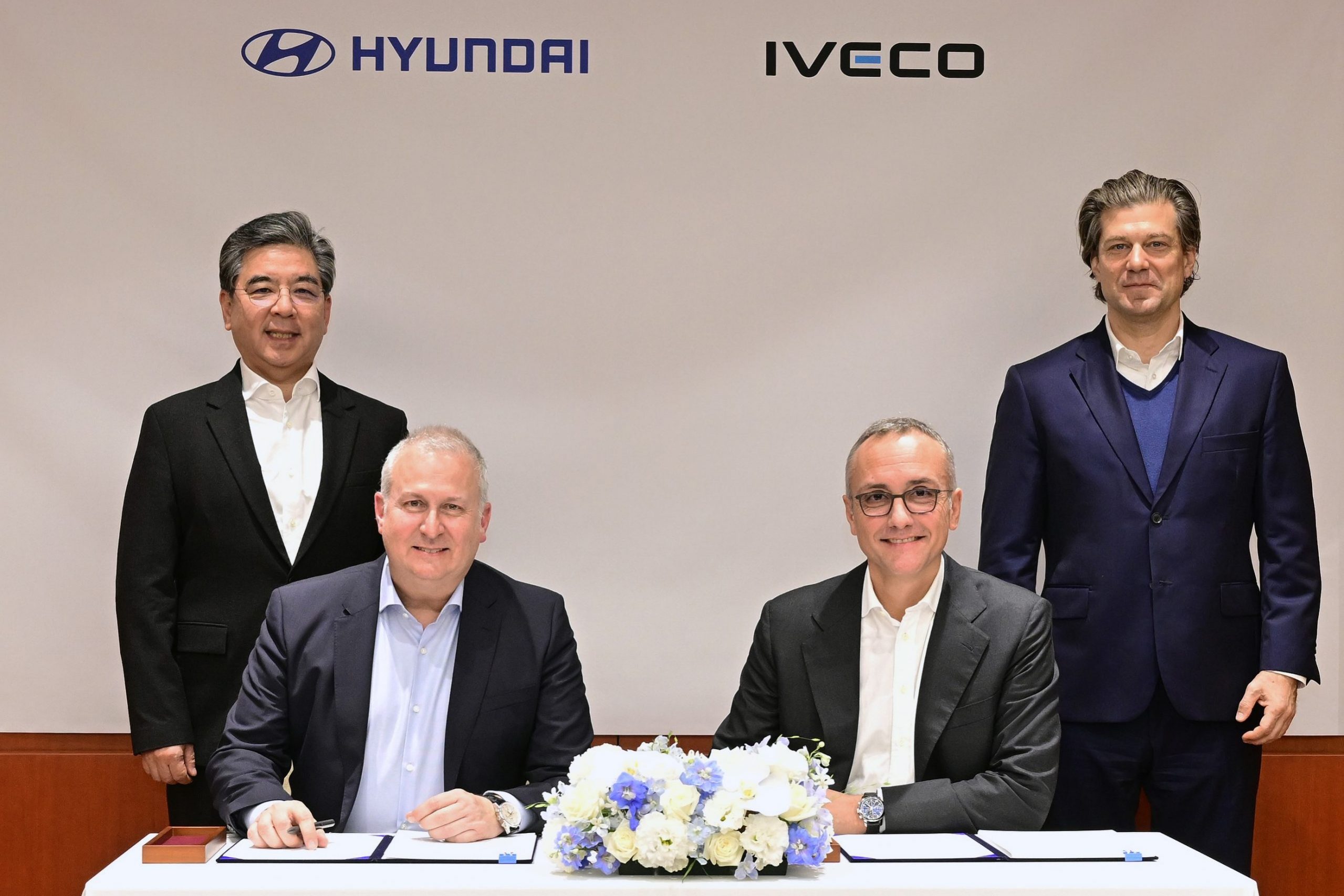 Hyundai liefert vollelektrisches leichtes Nutzfahrzeug an Iveco in Europa. Das Bild zeigt das Führungspersonal beider Häuser bei der feierlichen Unterschrift. Zwei Männer sitzen an einem Tisch, vor sich die Vertragsunterlagen und zwischen ihnen ein Strauß Blumen. Hinter ihnen stehen zwei weitere Männer.