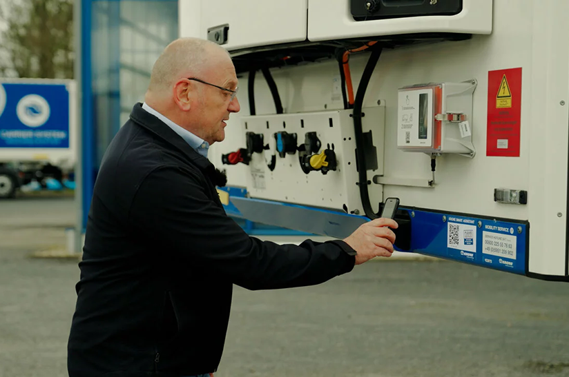 Krone_Smart_Assistant_web Das Bild zeigt einen Mann, der hinter einem LKW steht und mit seinem Smartphone einen QR-Code scannt. Er nutzt eine neue Lösung von Krone.
