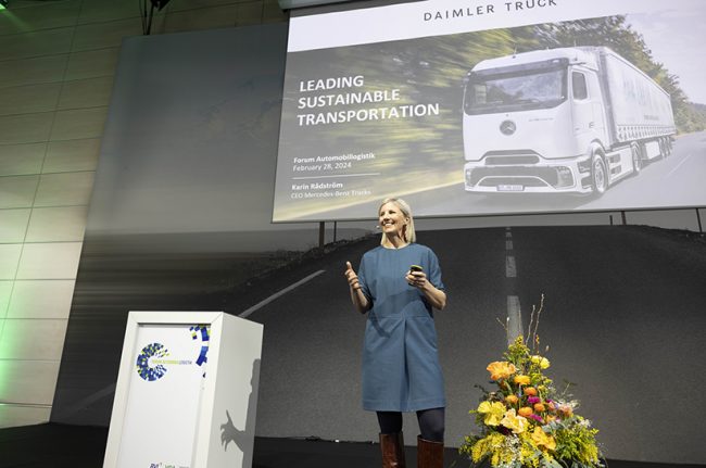 Das Bild zeigt eine weiße Frau, die auf einer Bühne steht. Im Hintergrund ist eine Leinwand mit einem Truck zu sehen. Was ein Wettbewerbsvorteil ist, war auch Thema der Veranstaltung.