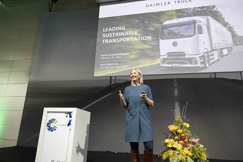BVL_VDA_Forum_automobillogisti_web Das Bild zeigt eine weiße Frau, die auf einer Bühne steht. Im Hintergrund ist eine Leinwand mit einem Truck zu sehen. Was ein Wettbewerbsvorteil ist, war auch Thema der Veranstaltung.