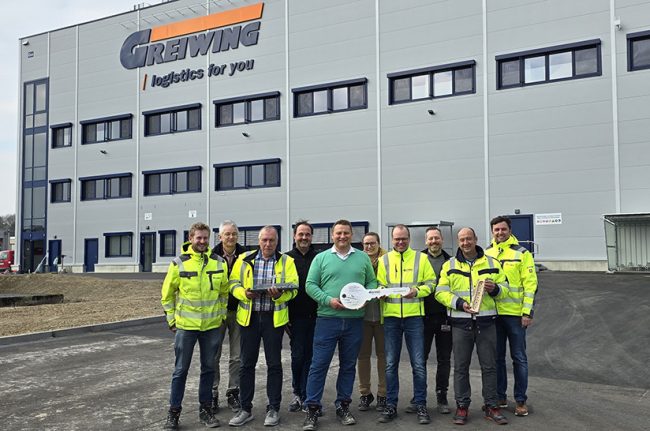 Das Bild zeigt Mitarbeiter des Logistikunternehmens Greiwing mit einem überdimensionalem Schlüssel Im Hintergrund ist das neu eröffnete Logistikzentrum zu sehen.