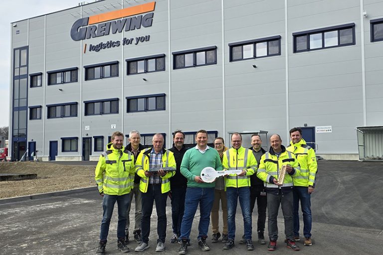 GREIWING Rheinmünster (web) Das Bild zeigt Mitarbeiter des Logistikunternehmens Greiwing mit einem überdimensionalem Schlüssel Im Hintergrund ist das neu eröffnete Logistikzentrum zu sehen.
