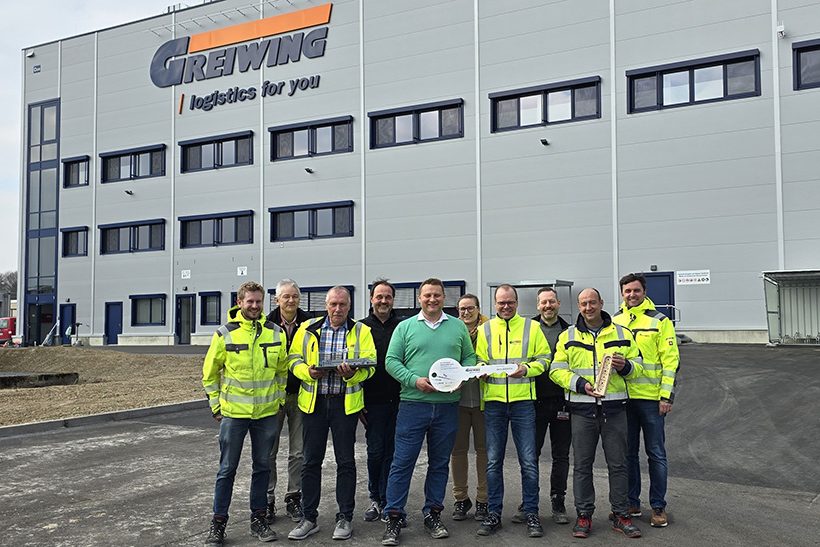 GREIWING Rheinmünster (web) Das Bild zeigt Mitarbeiter des Logistikunternehmens Greiwing mit einem überdimensionalem Schlüssel Im Hintergrund ist das neu eröffnete Logistikzentrum zu sehen.