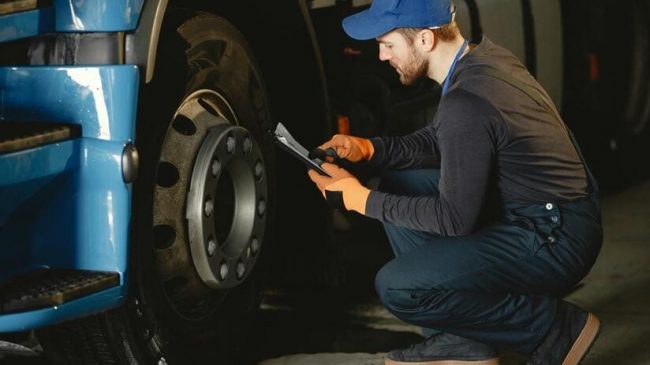 Reifendruckkontrollsysteme werden für neu zugelassene Nutzfahrzeuge Pflicht . Das Bild zeigt einen Mechaniker vor einem Lkw-Reifen- Er kniet uns hält eine Klemmmappe in der Hand.