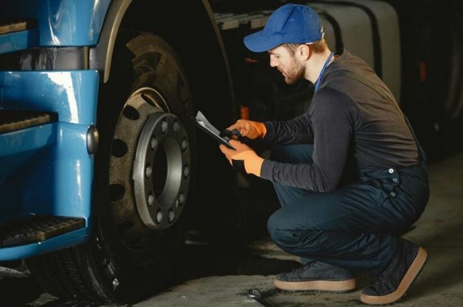 Reifendruckkontrollsysteme werden für neu zugelassene Nutzfahrzeuge Pflicht . Das Bild zeigt einen Mechaniker vor einem Lkw-Reifen- Er kniet uns hält eine Klemmmappe in der Hand.